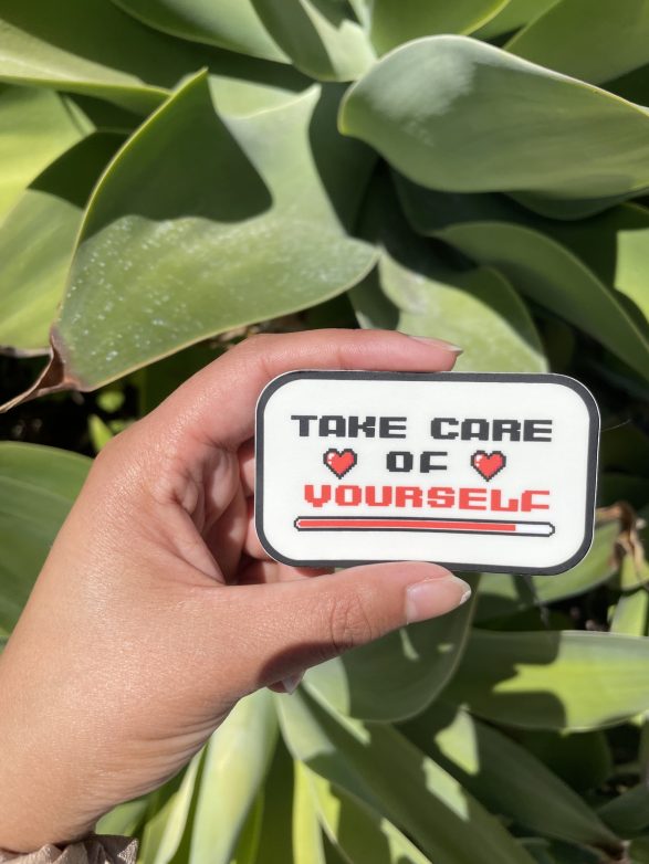 Aleenah's "Take Care of Yourself" sticker being held in front of some greenery