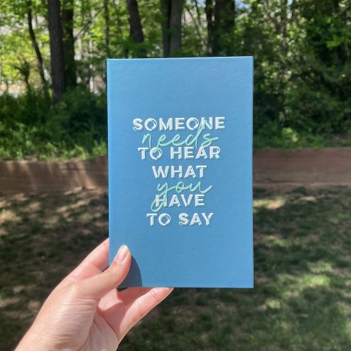 A hand holding up a teal notebook with the words "Someone needs to hear what you have say" written in graphic, block letters on the front