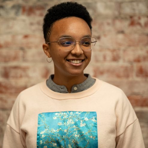 A young person with tan skin and short, black hair, wearing glasses, small gold hoop earrings, and a white pullover sweatshirt. They are smiling and looking just to the right fo the camera.
