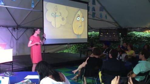 Hanna speaking on a stage wearing pink with a drawing slide deck behind her